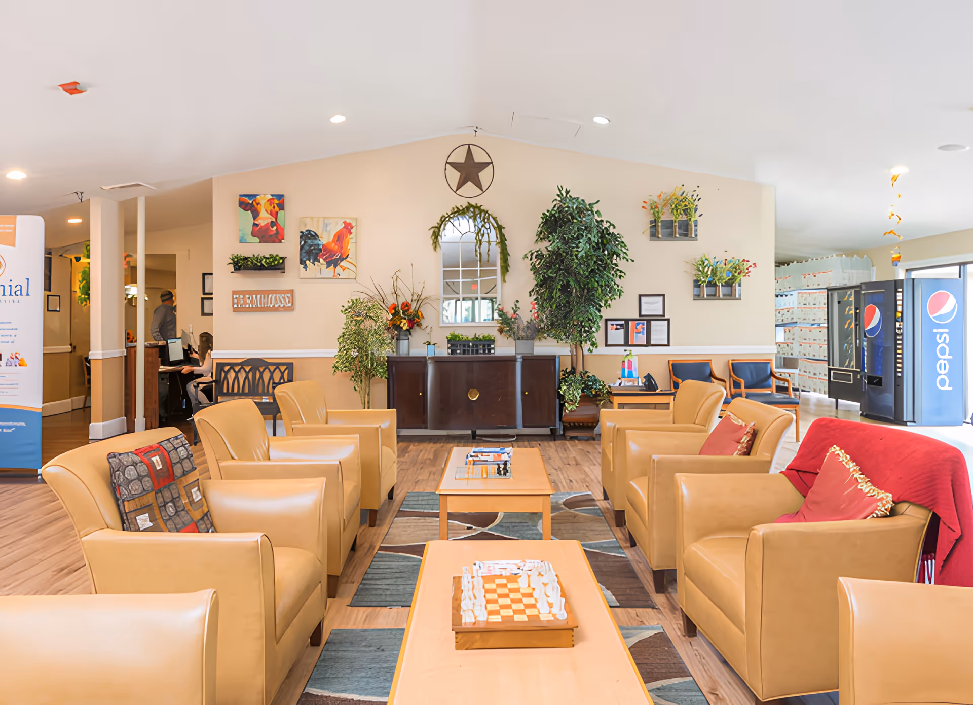 A bright and welcoming living room area in Colonial Assisted Living at Miami, featuring tan leather armchairs arranged around wooden coffee tables with a chessboard on one table. The room has wooden flooring, decorative plants, colorful wall art including a cow and rooster painting, a mirror with greenery, and a star decoration above it. In the background, there is a Pepsi vending machine and a reception area with staff working.
