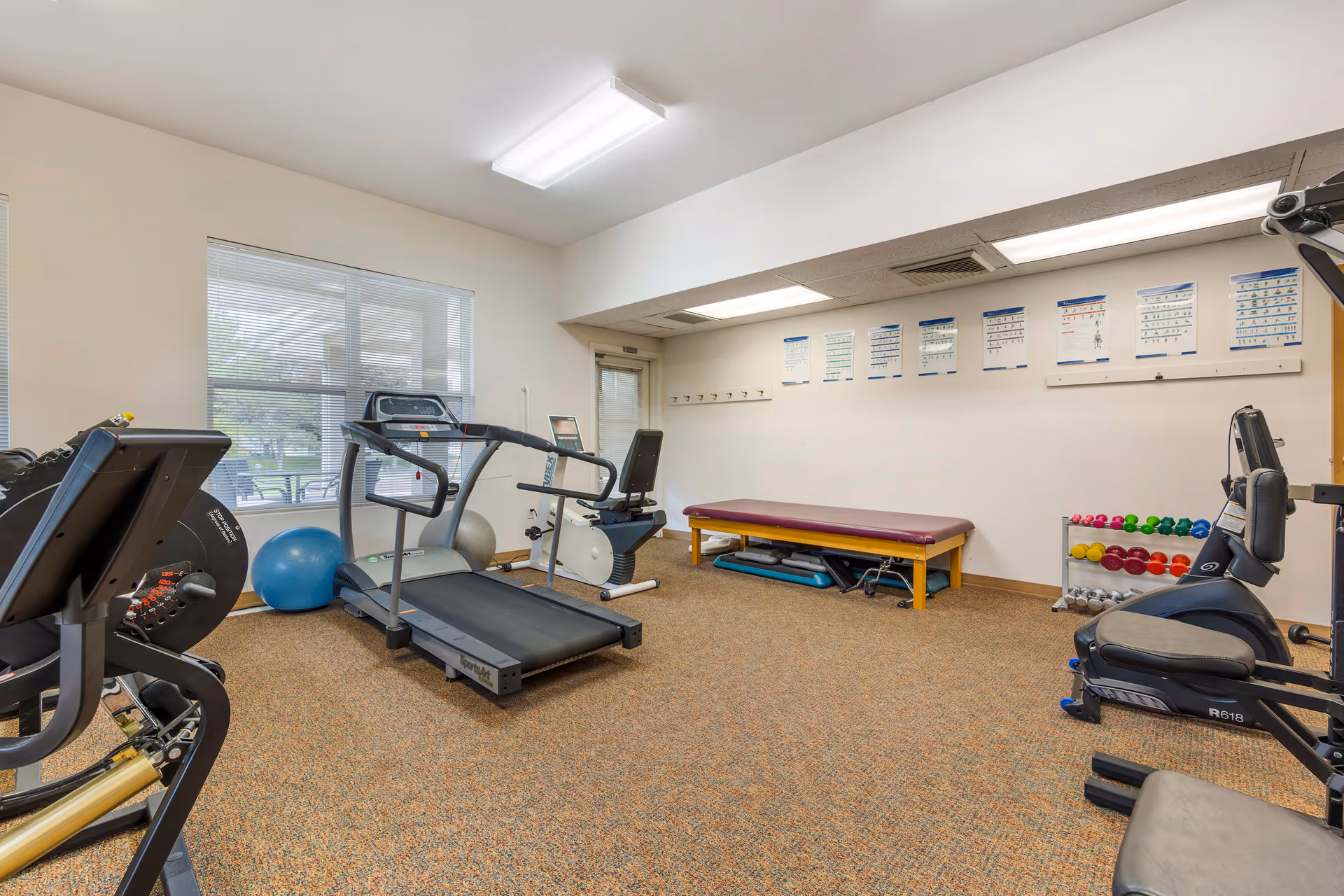 A fitness room with exercise equipment including a treadmill, stationary bike, weight machine, and a rack of colorful dumbbells. There is a padded bench against the wall with exercise charts displayed above it. The room has carpeted flooring, a window with blinds, and fluorescent ceiling lights.