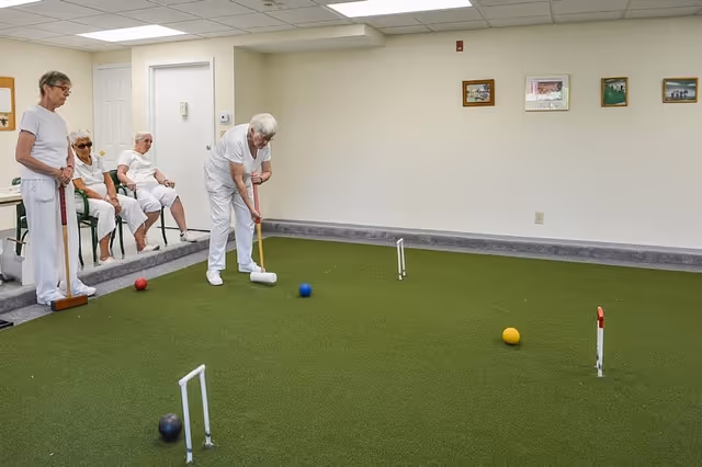Four elderly women in white clothing playing indoor croquet on a green carpeted court inside a room with beige walls and framed pictures. Three women are seated on chairs while one woman is standing and preparing to hit a croquet ball with a mallet.