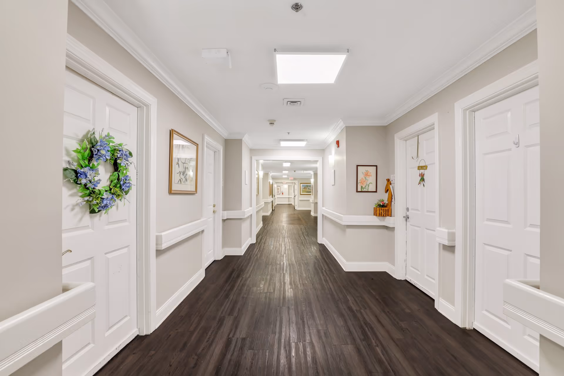 Long, well-lit interior hallway of an assisted living facility with doors on both sides and dark wood flooring.