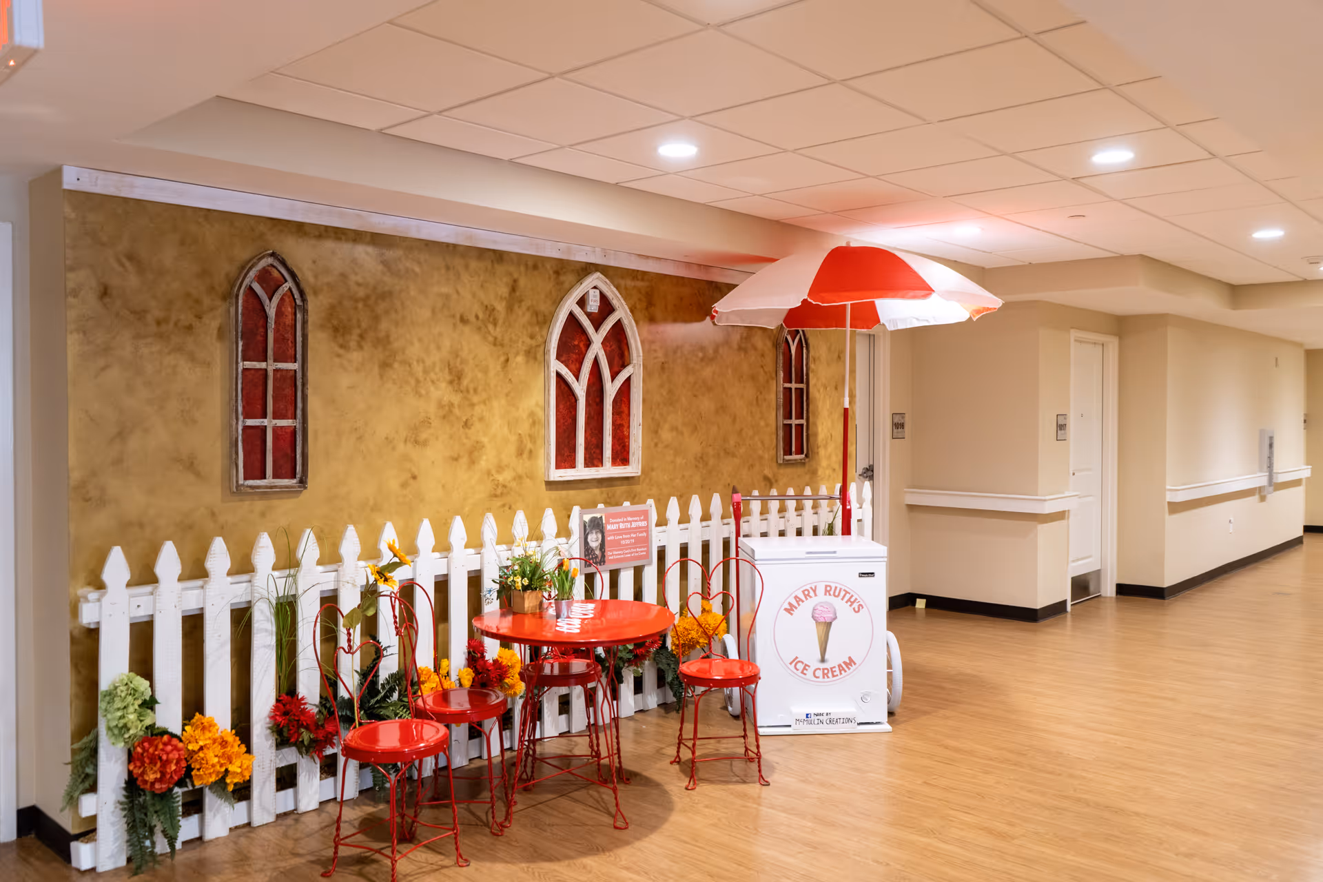 Indoor hallway area with a decorative white picket fence, red metal table and chairs, colorful flowers, and a white ice cream cart with a red and white umbrella labeled 'Mary Ruth's Ice Cream'. The wall behind has three arched window decorations with red panes.