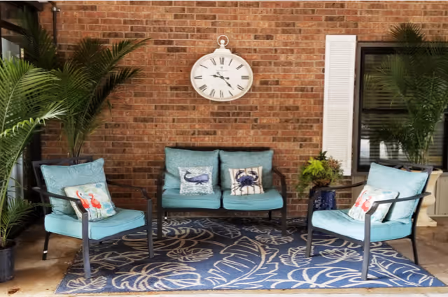 Outdoor seating area with two single chairs and one loveseat with teal cushions and decorative pillows featuring sea creatures, placed on a blue patterned rug against a brick wall with a white clock hanging above. Two potted palm plants flank the seating area, and a window with white shutters is visible on the right.