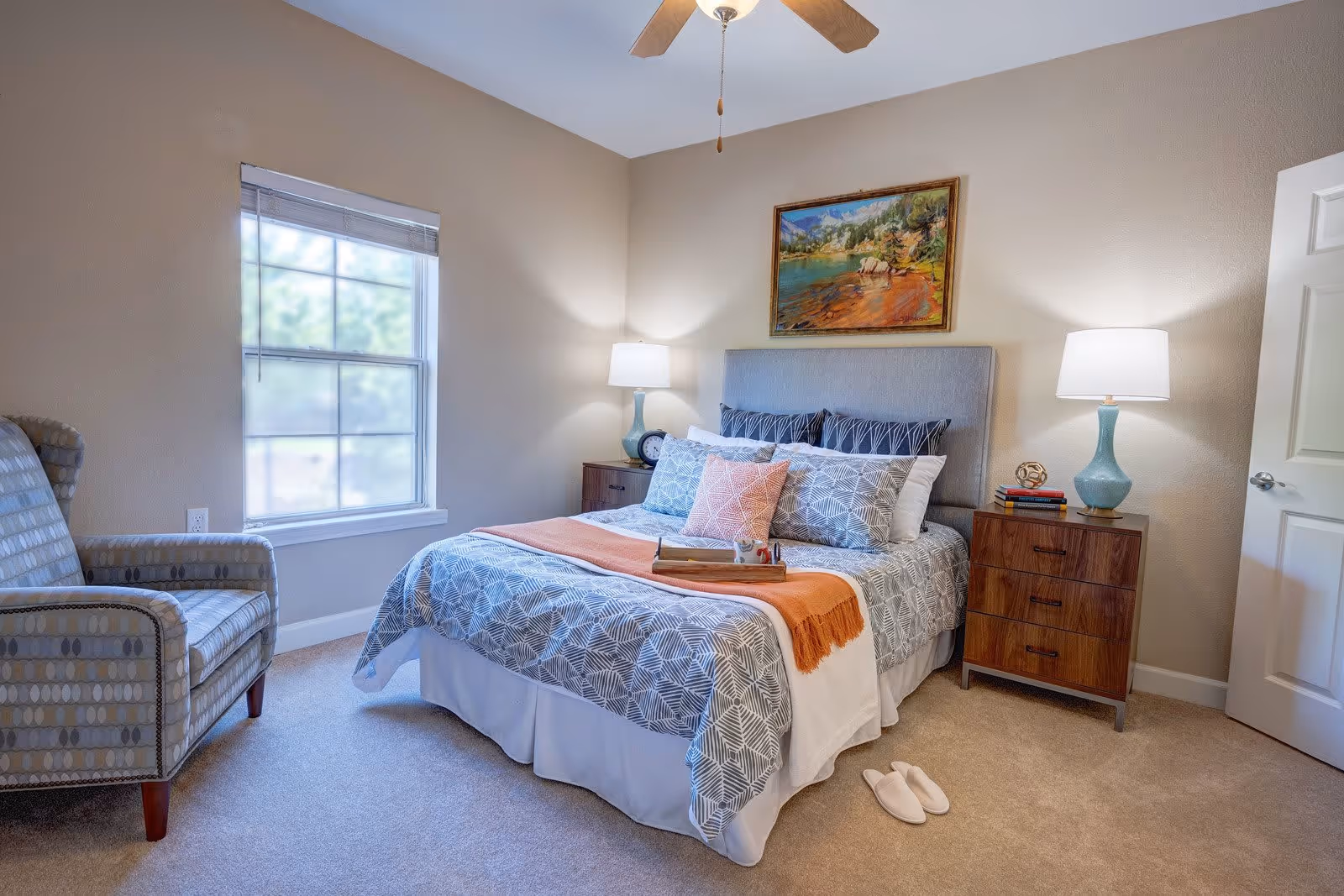 A cozy bedroom with a neatly made bed featuring patterned bedding and multiple pillows. There is an orange throw blanket at the foot of the bed and a wooden tray on top. On either side of the bed are wooden nightstands with blue lamps and decorative items. A comfortable patterned armchair is placed near a window with blinds. A ceiling fan is visible above, and a colorful landscape painting hangs on the wall above the bed. A pair of white slippers is on the carpeted floor next to the bed.