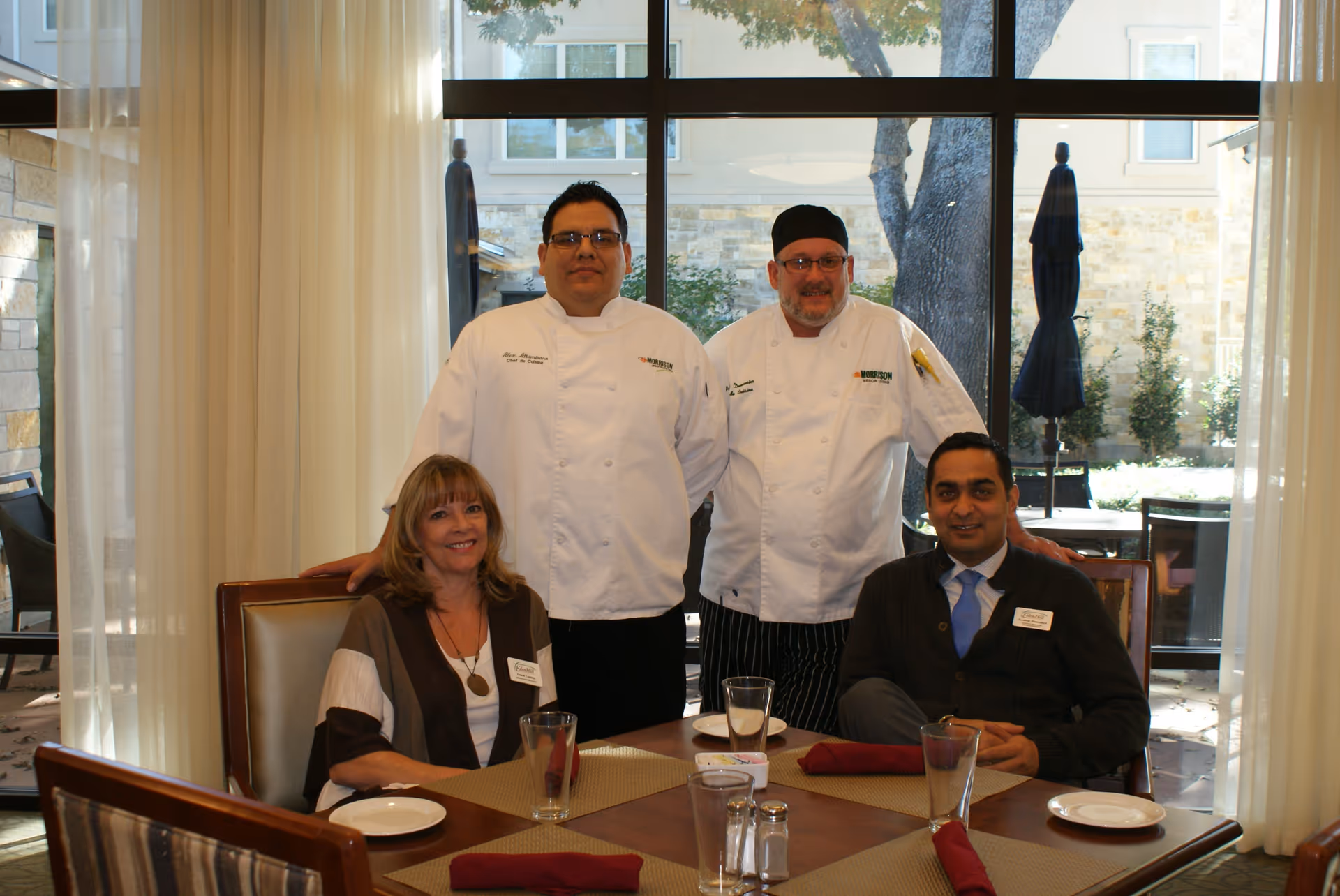 Two chefs and two staff members pose around a dining table in a bright dining room with large windows.