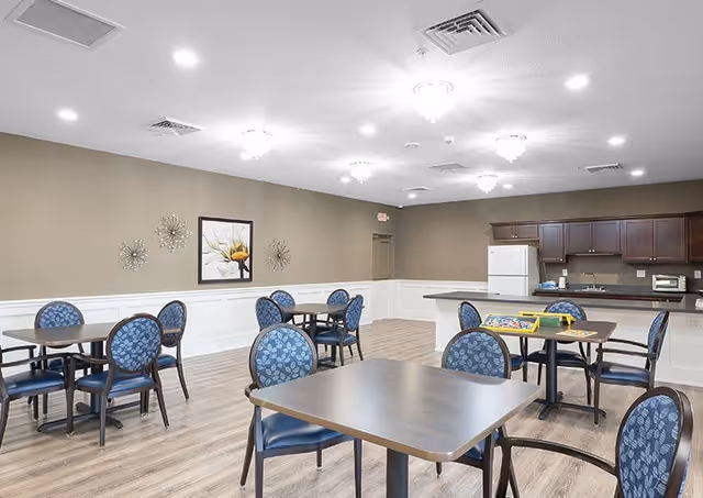 A bright and clean dining area with several square tables and blue cushioned chairs. The room has light wood flooring, beige walls with white wainscoting, and ceiling lights. There is a kitchen area in the background with dark wood cabinets, a white refrigerator, and countertop appliances. A framed flower artwork and decorative wall pieces are visible on the wall.