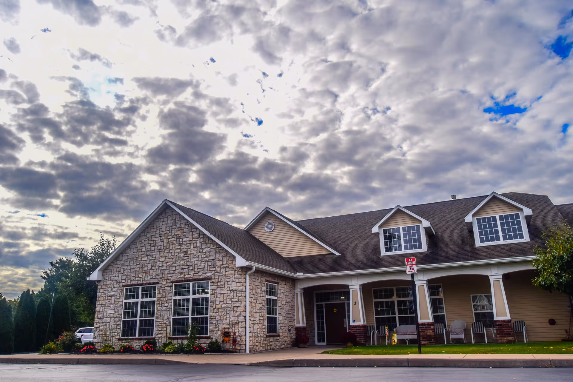 Exterior view of a senior living community building with stone and siding facade, multiple windows, a covered porch with chairs, and a cloudy sky overhead.