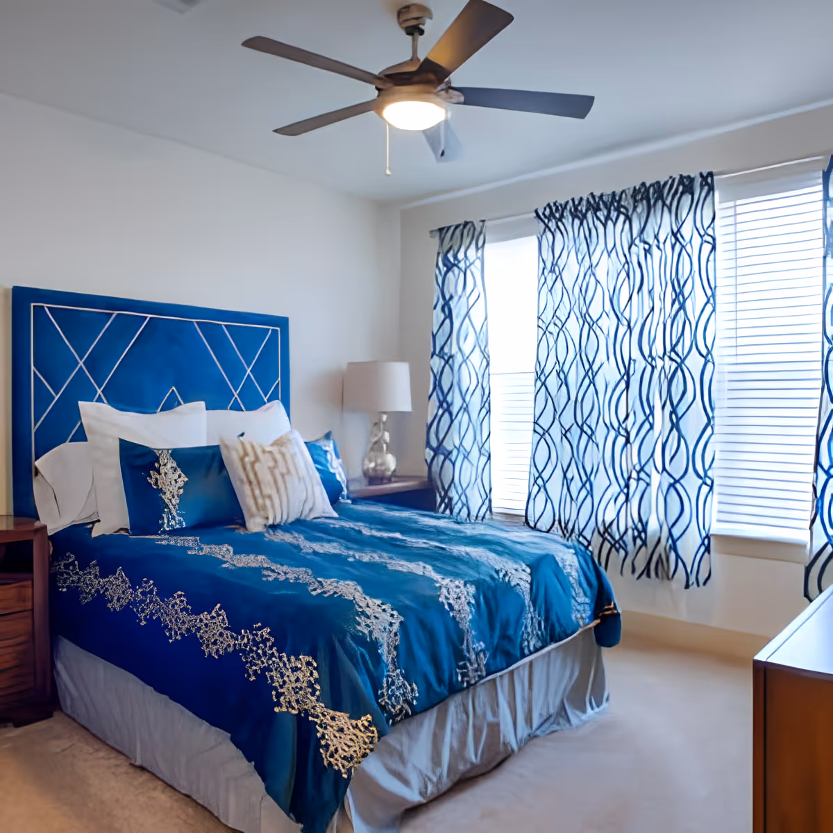 A bedroom with a bed featuring a blue and gold patterned comforter and matching pillows. The bed has a blue headboard with a geometric design. There is a wooden nightstand with a lamp on the left side of the bed. The room has large windows with white blinds and blue patterned curtains. A ceiling fan with a light is mounted on the ceiling.