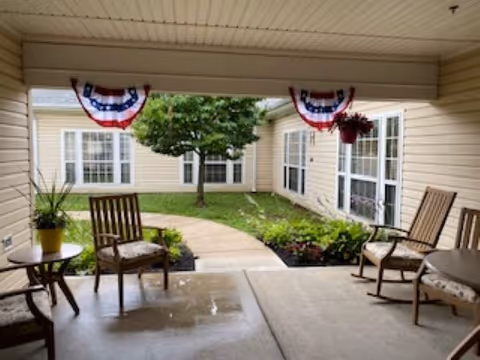 Covered patio area with wooden chairs and small tables, overlooking a courtyard with a tree and garden beds. The building has beige siding and multiple windows. Red, white, and blue bunting decorations hang from the patio ceiling.