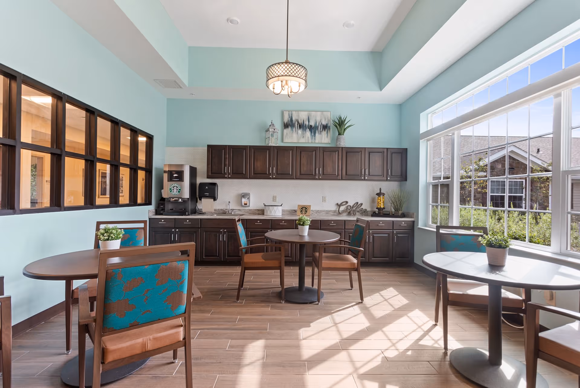 A bright room with light blue walls and large windows letting in natural light. The room features three round tables with wooden chairs that have teal and brown floral upholstery. Along the back wall are dark wood cabinets with a countertop, a coffee machine, and decorative items including a plant and a sign that says 'Coffee'. A modern ceiling light fixture hangs from the ceiling.