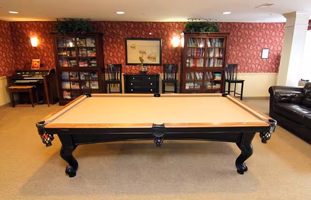 Pool table centered in a cozy common room with bookshelves, a piano, and seating.