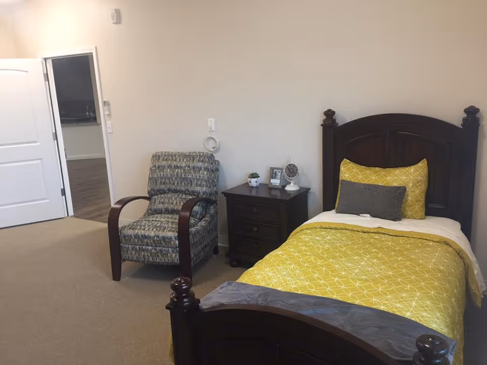 Single bedroom with a dark wood twin bed dressed in yellow bedding, a nightstand with decor, and an upholstered armchair.