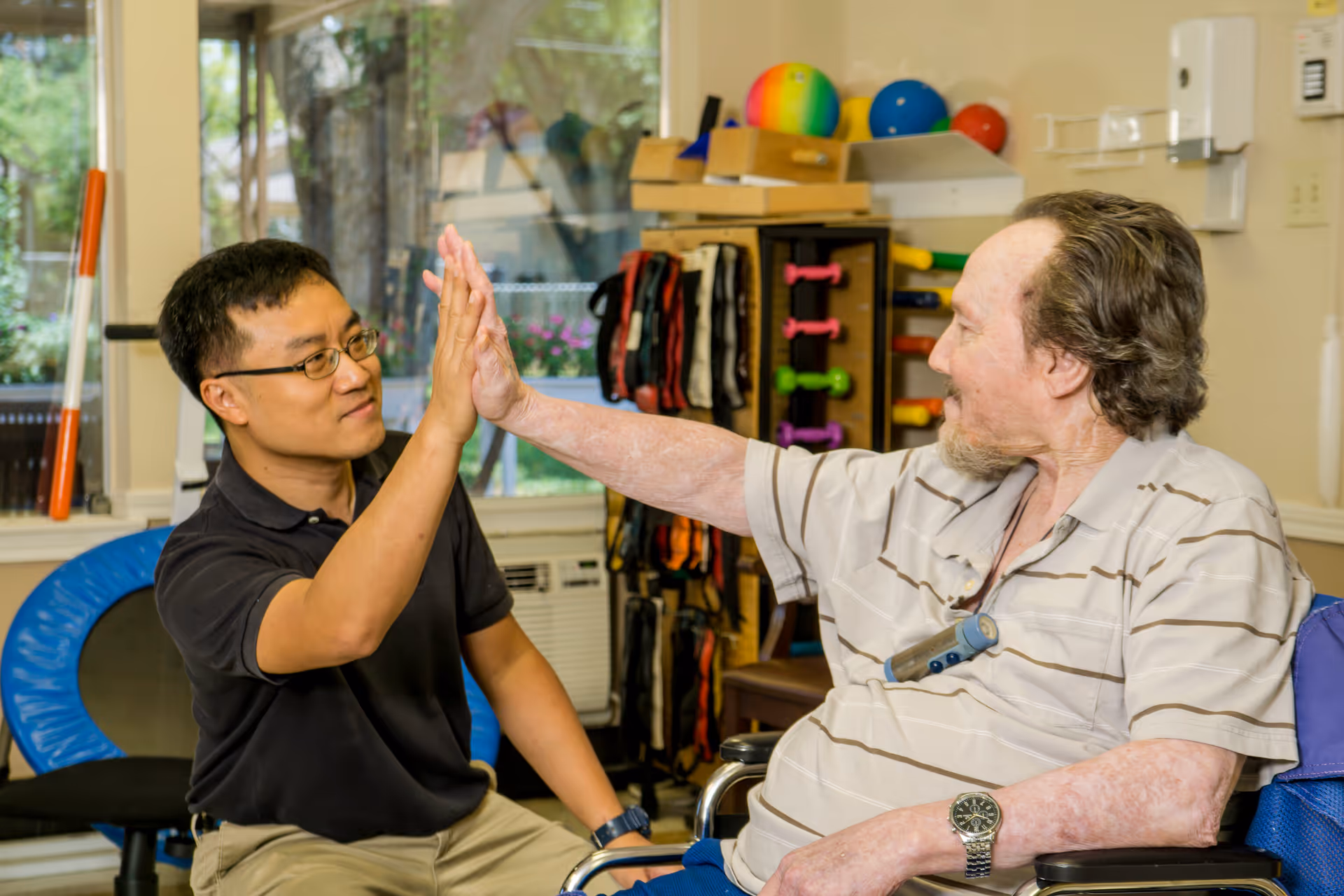 A man in a black shirt and glasses gives a high-five to an elderly man in a wheelchair wearing a striped polo shirt inside a room with exercise equipment and a window showing greenery outside.