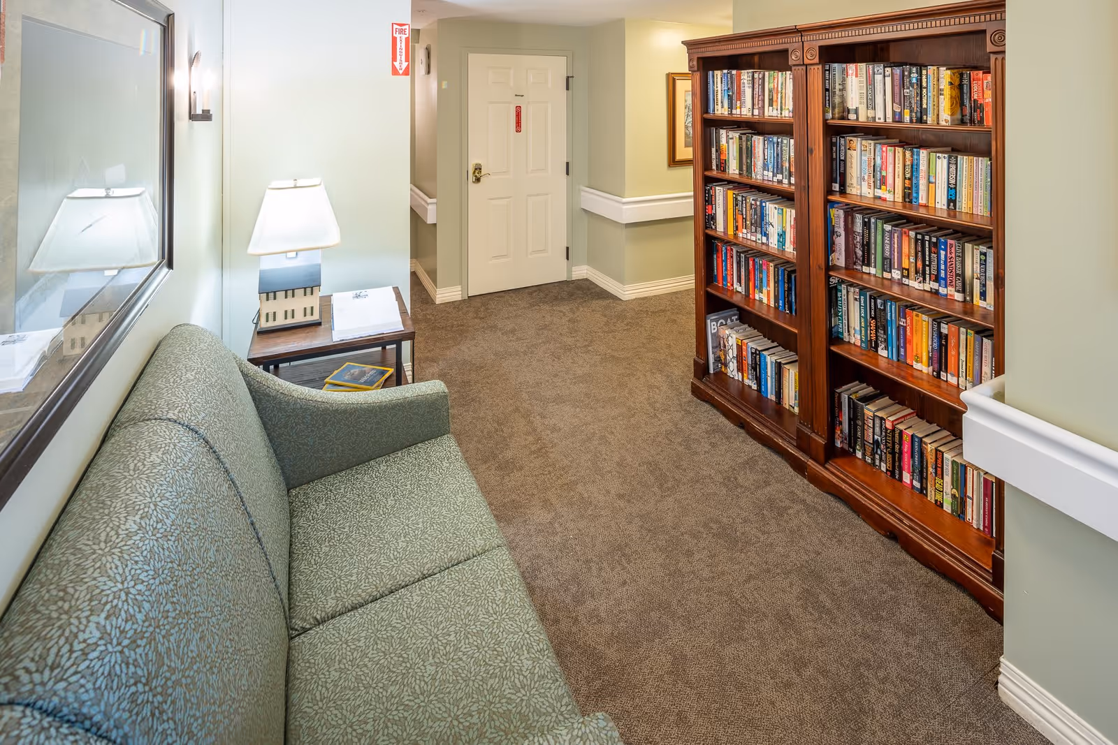 A cozy hallway area in a senior living facility featuring a green patterned couch, a wooden side table with a lamp and books, and a large wooden bookshelf filled with books. The walls are painted light green with white trim, and there is a door at the end of the hallway.