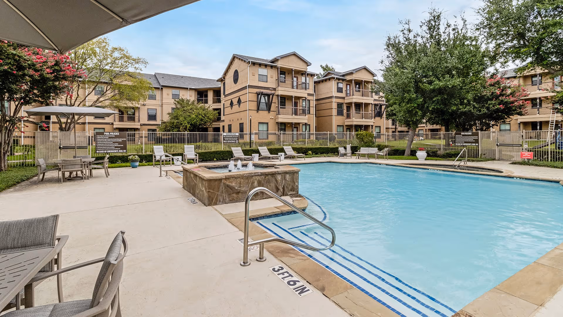 Outdoor swimming pool area at a senior living facility with lounge chairs, tables with umbrellas, and a hot tub. Surrounding the pool are three-story beige residential buildings and trees under a partly cloudy sky.