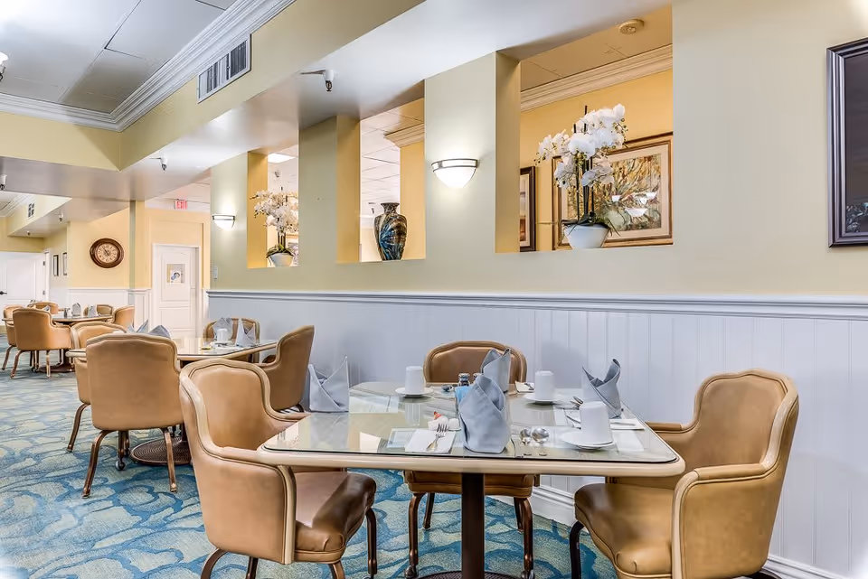 A dining area in a senior living facility with several tables set with napkins, cups, and utensils. The room has beige walls with white wainscoting, decorative wall niches with vases and flowers, and comfortable tan chairs around glass-top tables. The carpet has a blue pattern, and there is a clock on the far wall.