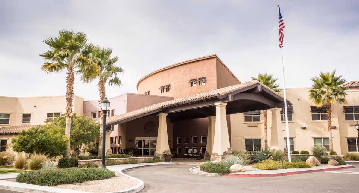 Front exterior view of Solana at The Park Assisted Living Community building with a covered entrance, palm trees, landscaping, and an American flag on a flagpole.