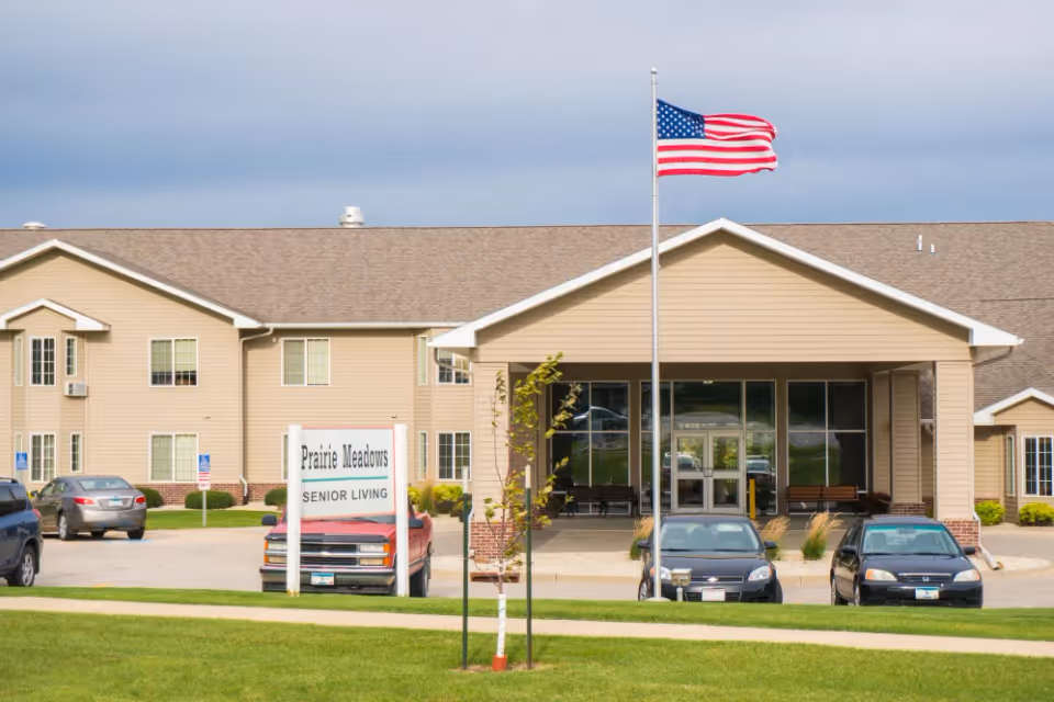 Front entrance of Prairie Meadows Senior Living with an American flag flying, a sign and several parked cars.
