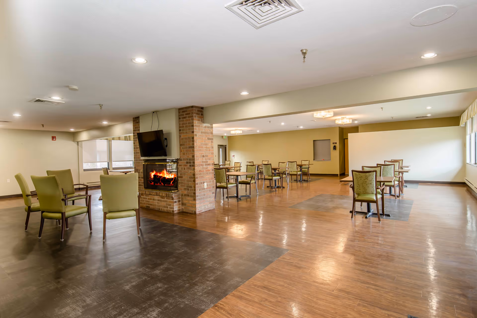 A spacious common area with multiple tables and chairs arranged for seating. There is a brick fireplace with a TV mounted above it on one side of the room. The floor has a mix of wood and dark tile sections, and the room is well-lit with ceiling lights and natural light from windows.