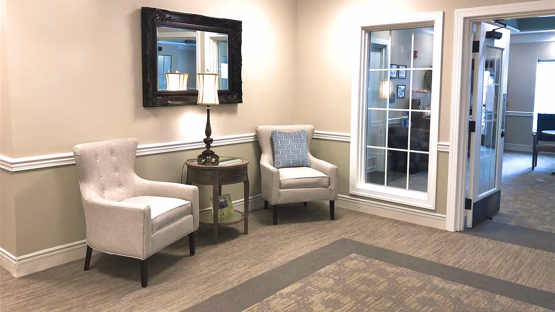 A cozy seating area in a facility with two beige upholstered armchairs separated by a small round wooden table with a lamp on it. A large ornate mirror hangs on the wall above the table. To the right, there is a window with white trim and a door leading to another room with office furniture visible inside.