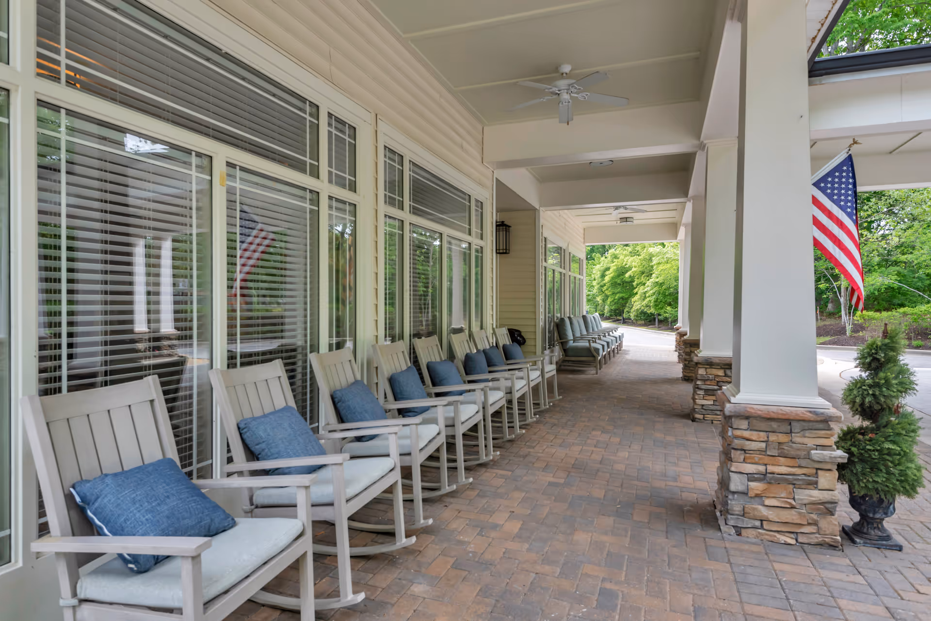 Covered front porch with a row of cushioned rocking chairs along windows and an American flag by the entrance.