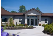 Front entrance of a single-story nursing and rehabilitation building with glass double doors, a paved circular driveway, and landscaped planting beds.