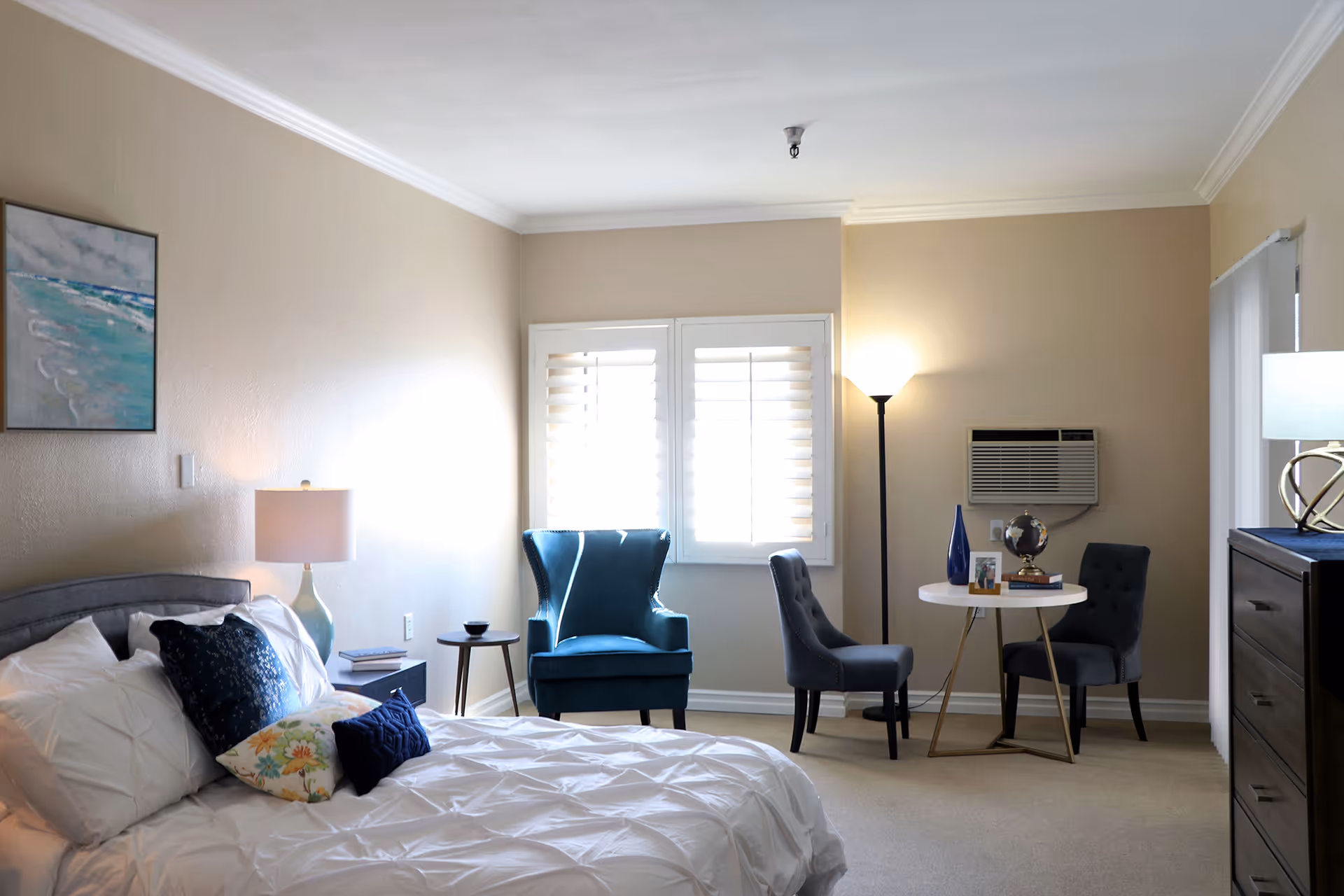A well-lit bedroom with a neatly made bed featuring white bedding and decorative pillows. To the left of the bed is a nightstand with a lamp and books. On the far wall, there is a window with white shutters, a blue armchair, and two gray chairs around a small round table with decorative items. A floor lamp and an air conditioning unit are also visible. A dresser with a lamp is on the right side of the room, and a painting hangs on the left wall.