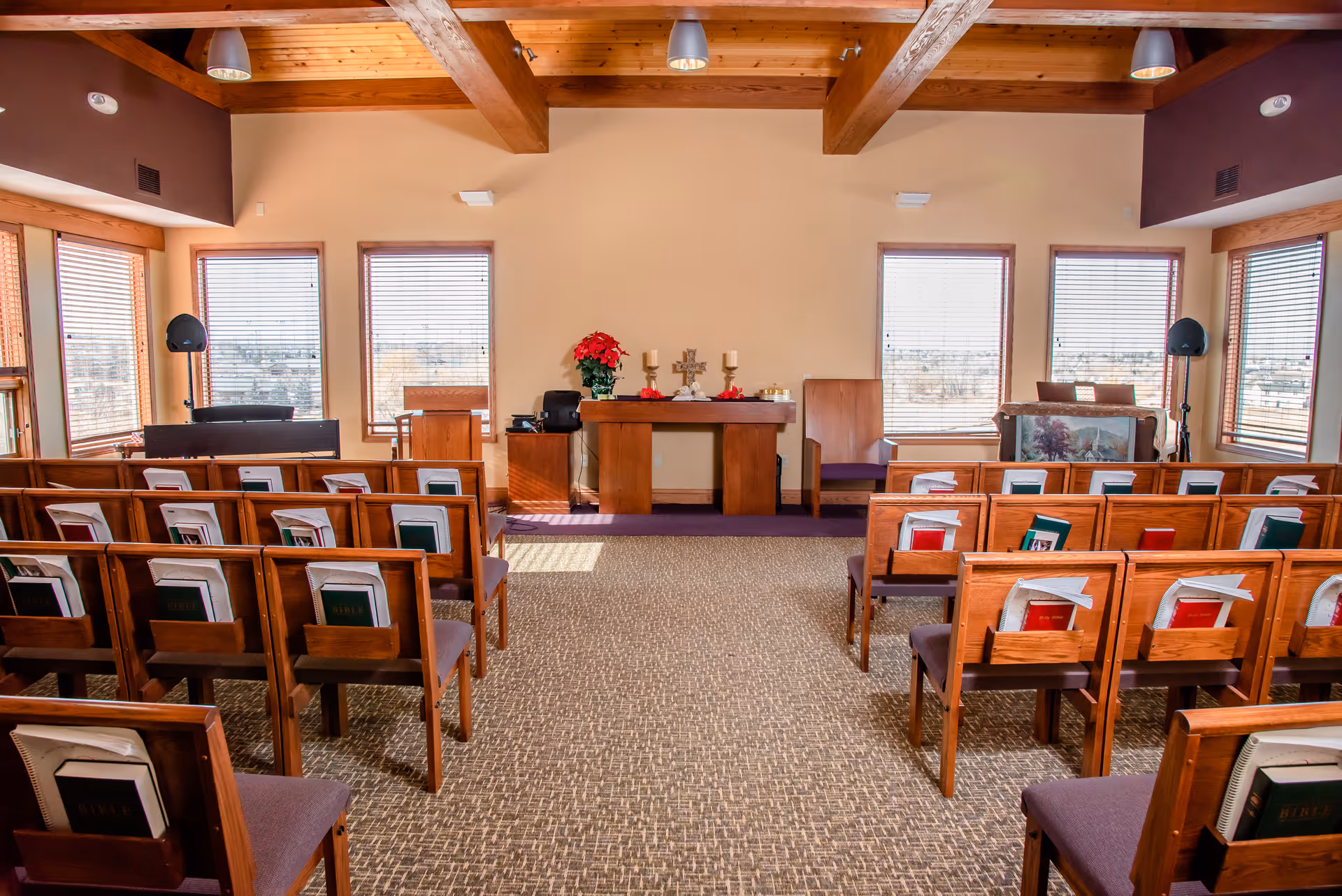 Interior view of a small chapel or worship room with wooden pews arranged in rows facing a wooden altar. The altar has candles, a cross, and a poinsettia plant. Large windows with blinds let in natural light, and the ceiling features exposed wooden beams.