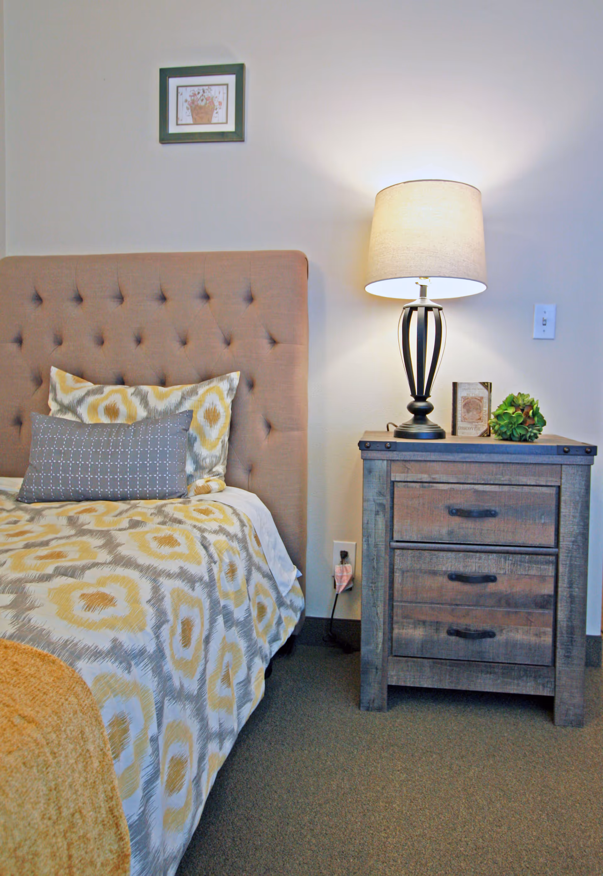 Tufted bed with patterned bedding and pillows next to a wooden nightstand topped with a lamp and small decor.