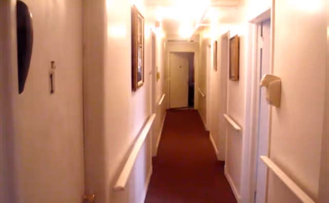 Narrow carpeted corridor with handrails, doors and framed artwork along the walls.