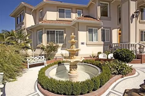 Exterior view of a two-story beige residential building with a tiled roof, featuring a circular water fountain surrounded by neatly trimmed bushes and a paved walkway. There are benches and various plants around the fountain area under a clear blue sky.