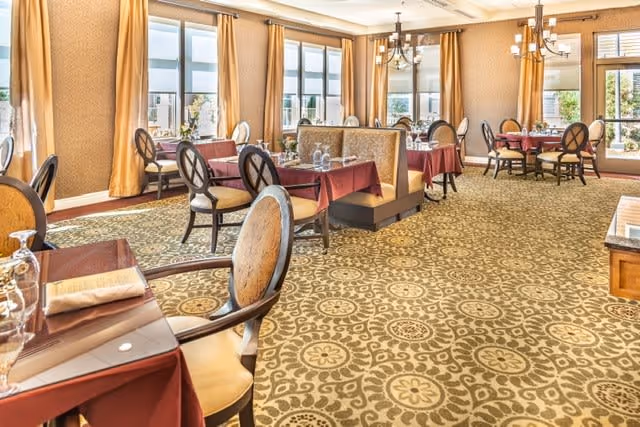 A spacious dining room with multiple tables covered in burgundy tablecloths, set with glassware and napkins. The room features large windows with golden curtains, patterned carpet, and elegant chandeliers hanging from the ceiling. Comfortable chairs and booth seating are arranged around the tables.