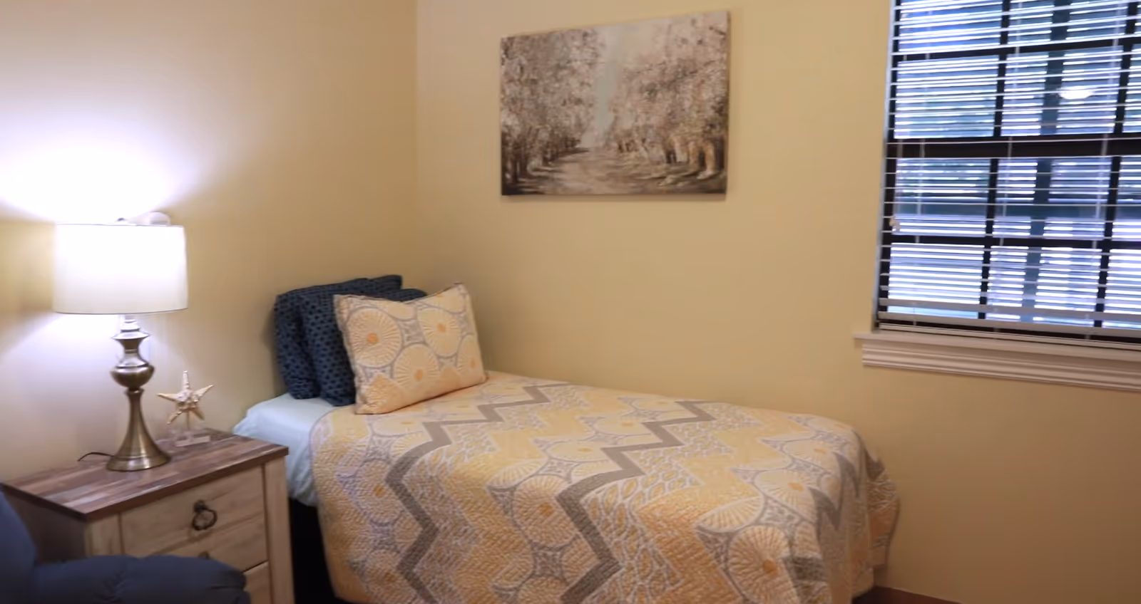 A small bedroom with a single bed covered in a yellow and gray patterned quilt and matching pillows. Next to the bed is a wooden nightstand with a silver lamp and a decorative starfish. A window with blinds is on the right wall, and a painting of a tree-lined path hangs above the bed.
