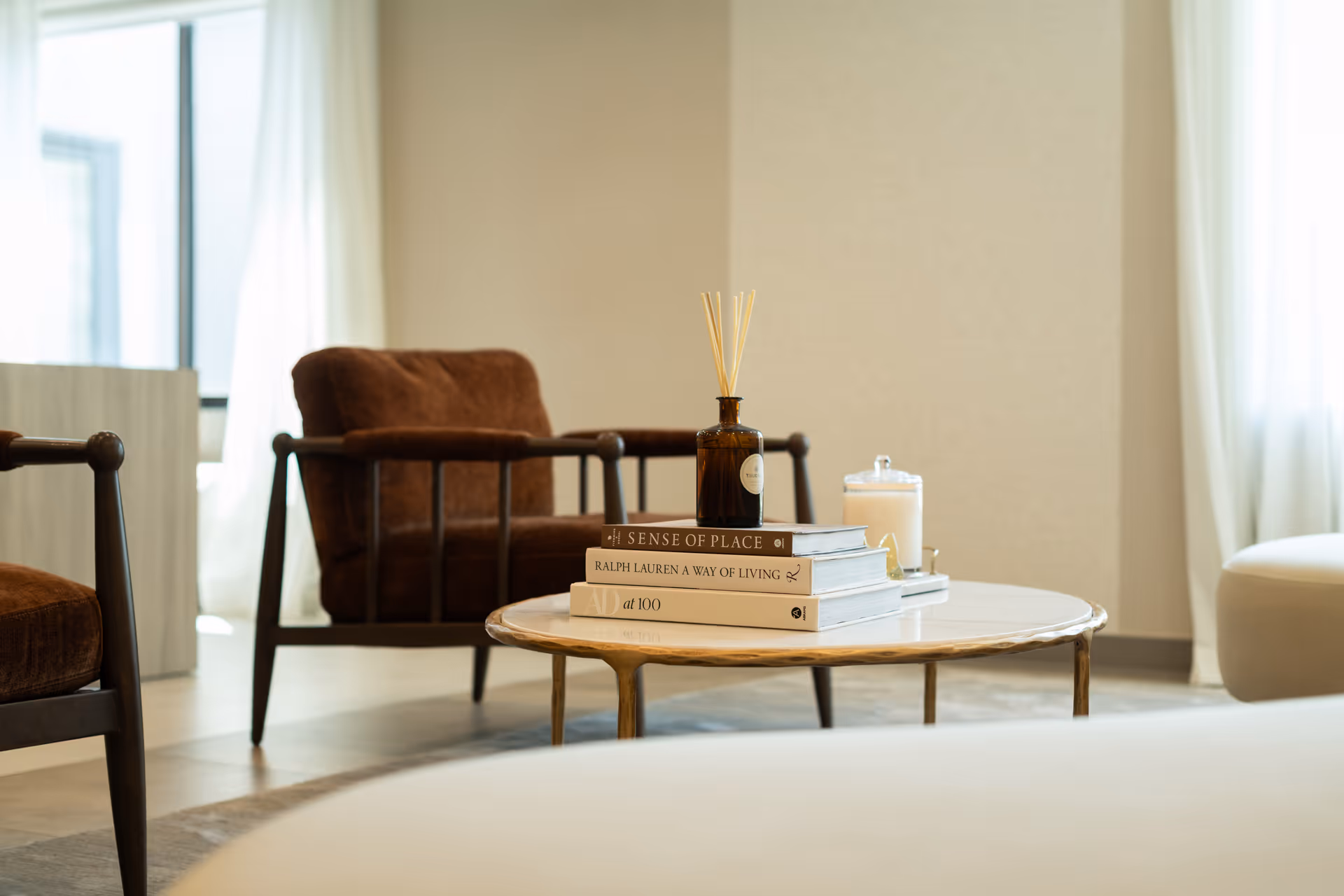 A cozy living room area with a round marble coffee table holding a stack of books, a brown glass diffuser bottle, and a white candle. In the background, there are two brown cushioned armchairs and light-colored curtains covering windows.