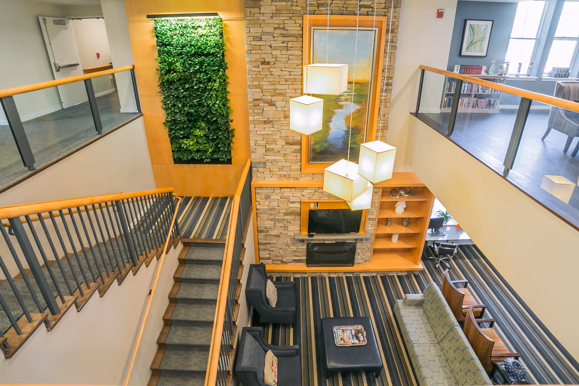 Interior view of a senior living facility lounge area with a staircase leading down to a seating area. The seating area includes a green patterned sofa, two dark armchairs, a black ottoman with magazines, and a stone fireplace with a mounted TV above it. There is a vertical green plant wall and a large framed painting above the fireplace. Modern pendant lights hang from the ceiling, and the upper level has a railing with glass panels and wooden handrails.