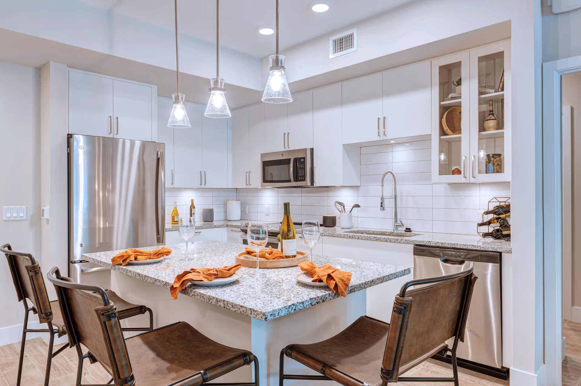 Modern kitchen with white cabinets, stainless steel refrigerator, microwave, and dishwasher. A granite island with four brown chairs is set with plates, orange napkins, and wine glasses. Three pendant lights hang above the island.