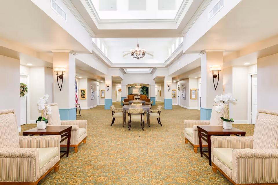 A spacious, well-lit common area in a senior living facility with high ceilings and a skylight. The room features beige walls with blue wainscoting, patterned carpet, and multiple seating arrangements including striped armchairs with side tables and a central table with chairs. Wall sconces provide additional lighting, and decorative plants and artwork adorn the space.