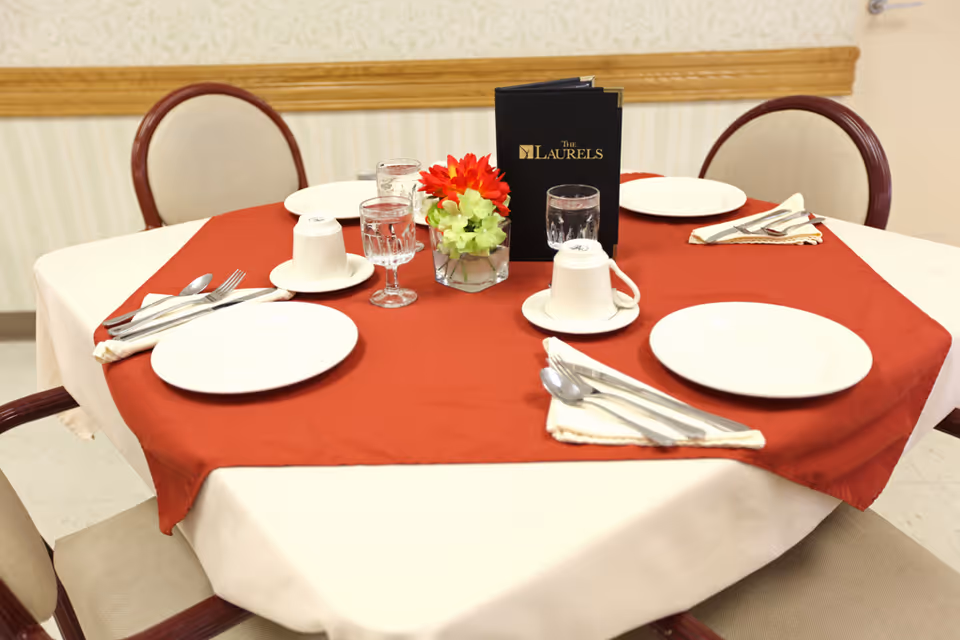 A dining table set for four with white plates, silverware wrapped in napkins, upside-down white cups on saucers, and glasses of water. The table has a red tablecloth over a white one, and a small flower arrangement with red and green flowers is in the center along with a black menu labeled 'The Laurels'. Four beige cushioned chairs with wooden frames surround the table.