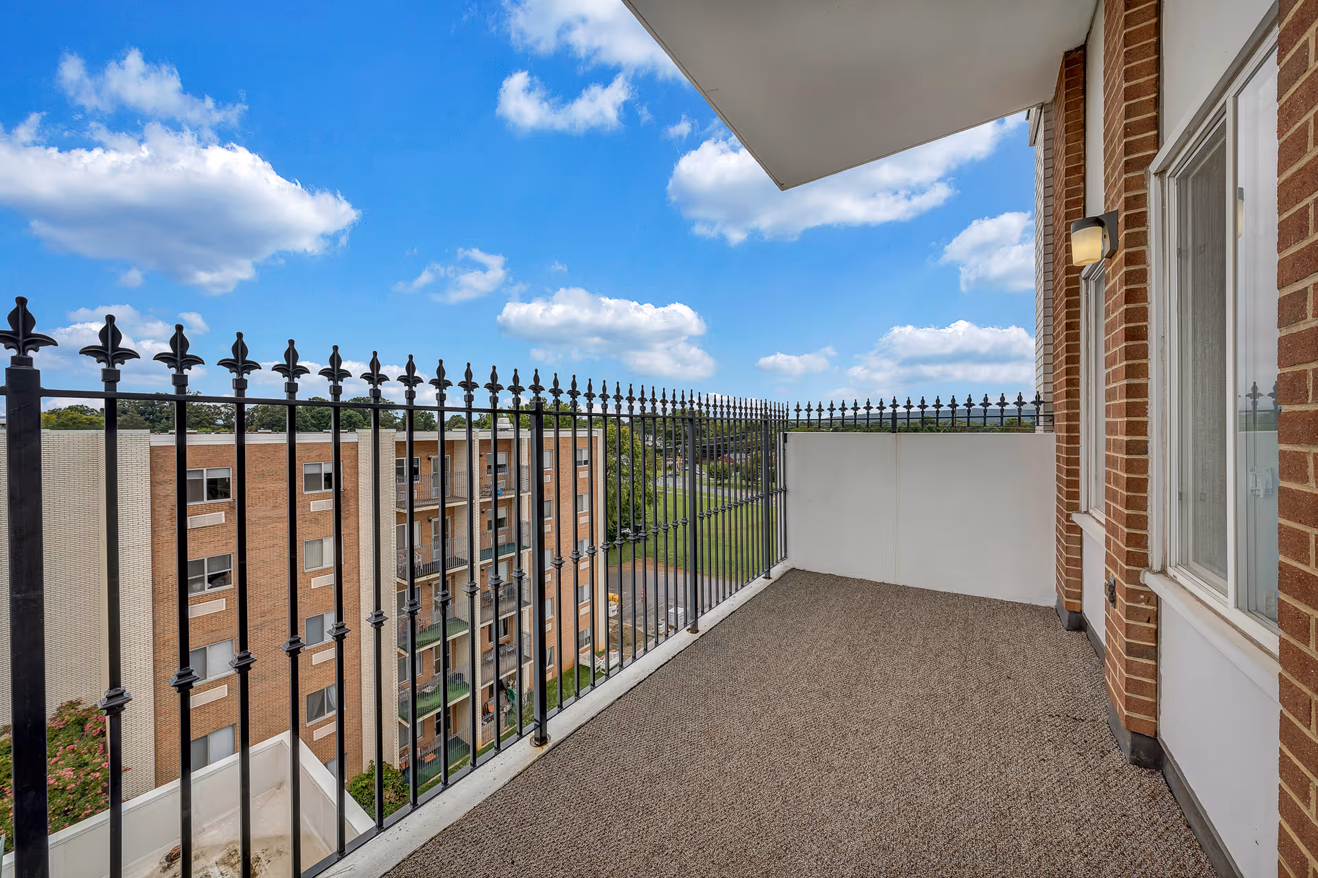 A balcony with a black wrought iron railing overlooking a neighboring brick building and green lawn under a blue sky with scattered clouds. The balcony has a carpeted floor and is attached to a brick wall with a window and an outdoor light fixture.
