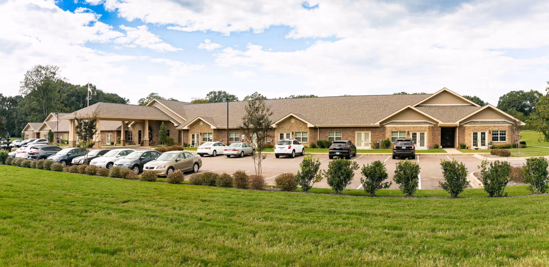 Exterior view of Hearthside Senior Living facility showing a single-story building with a large parking lot in front, several cars parked, green lawn and bushes in the foreground, and a partly cloudy sky above.