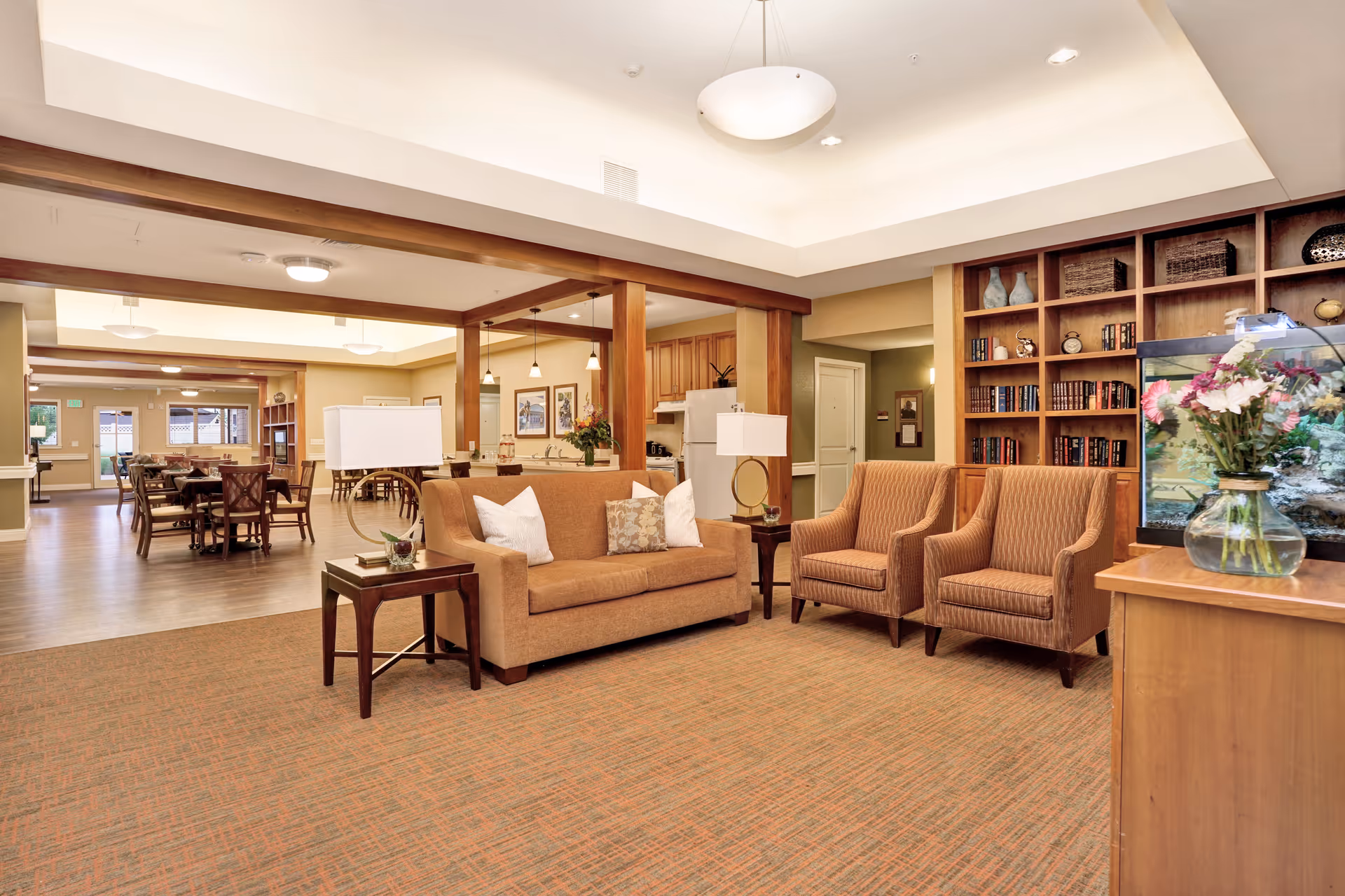 A cozy common area in a senior living facility featuring a brown sofa with white and patterned pillows, two matching armchairs, a side table with a lamp, a bookshelf filled with books and decorative items, and a fish tank with flowers on top. In the background, there is a dining area with tables and chairs, and a kitchen area with wooden cabinets and pendant lighting.