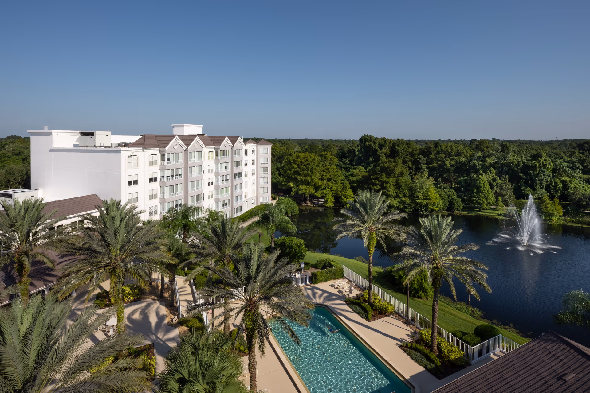 View of a multi-story senior living facility building with a swimming pool surrounded by palm trees in the foreground and a large pond with a fountain in the background under a clear blue sky.