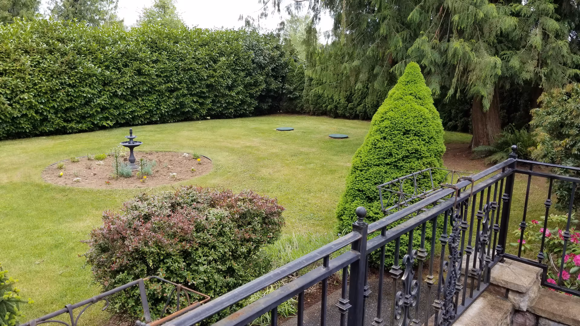 A well-maintained outdoor garden area with a circular flower bed containing a small black fountain. The garden is surrounded by dense green hedges and tall trees. In the foreground, there is a black wrought iron railing with some pink flowers visible on the right side.