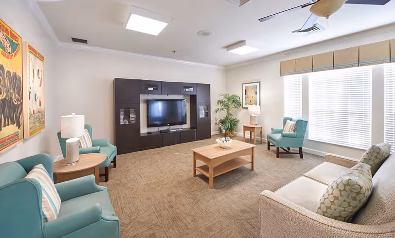 A bright and cozy living room with a beige sofa and two teal armchairs arranged around a wooden coffee table. There is a dark wood entertainment center with a flat-screen TV on the far wall. The room has large windows with white blinds and beige valances, two side tables with lamps, a potted plant, and framed artwork on the walls.