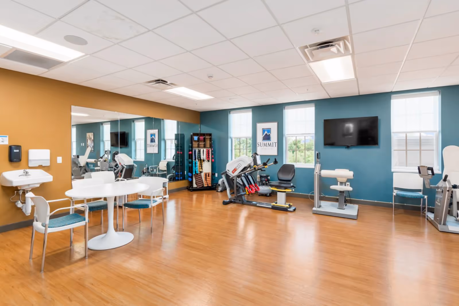 Bright therapy and fitness room with exercise equipment, a round table and chairs, wall mirrors, and a mounted TV.