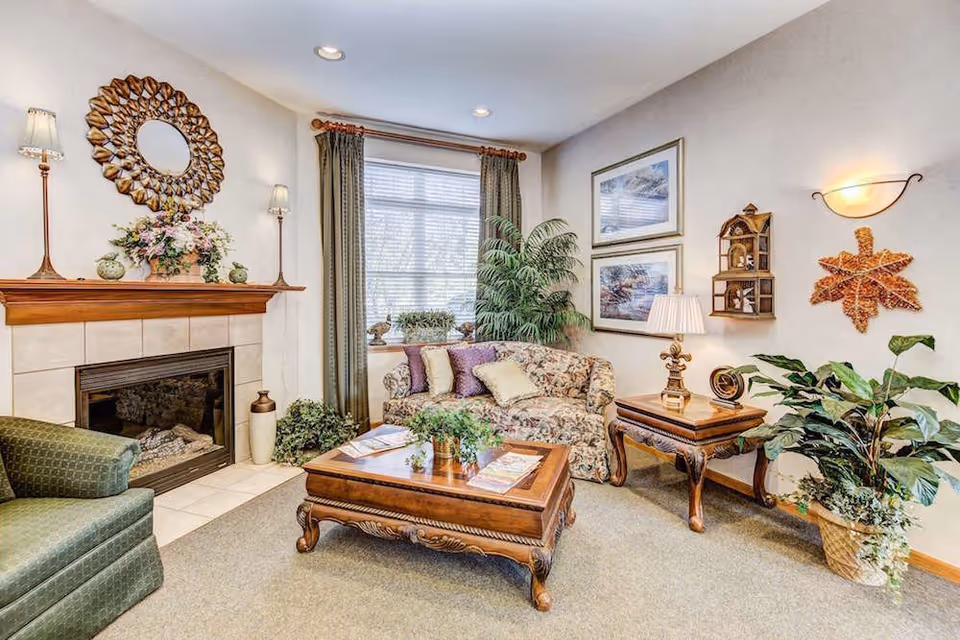Bright, traditionally furnished living room with a fireplace, floral sofa, wooden coffee table, and potted plants.