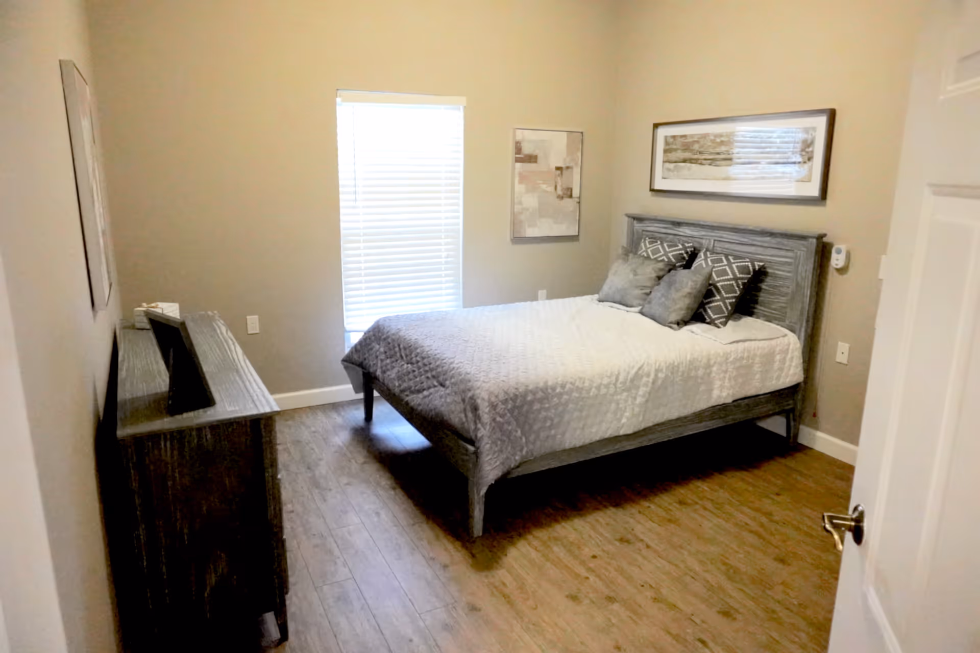 A bedroom with a wooden bed frame and a neatly made bed with gray and white bedding and pillows. There is a wooden dresser with a framed picture on top against the left wall. The room has light-colored walls, a window with blinds, two framed artworks on the walls, and wood flooring.