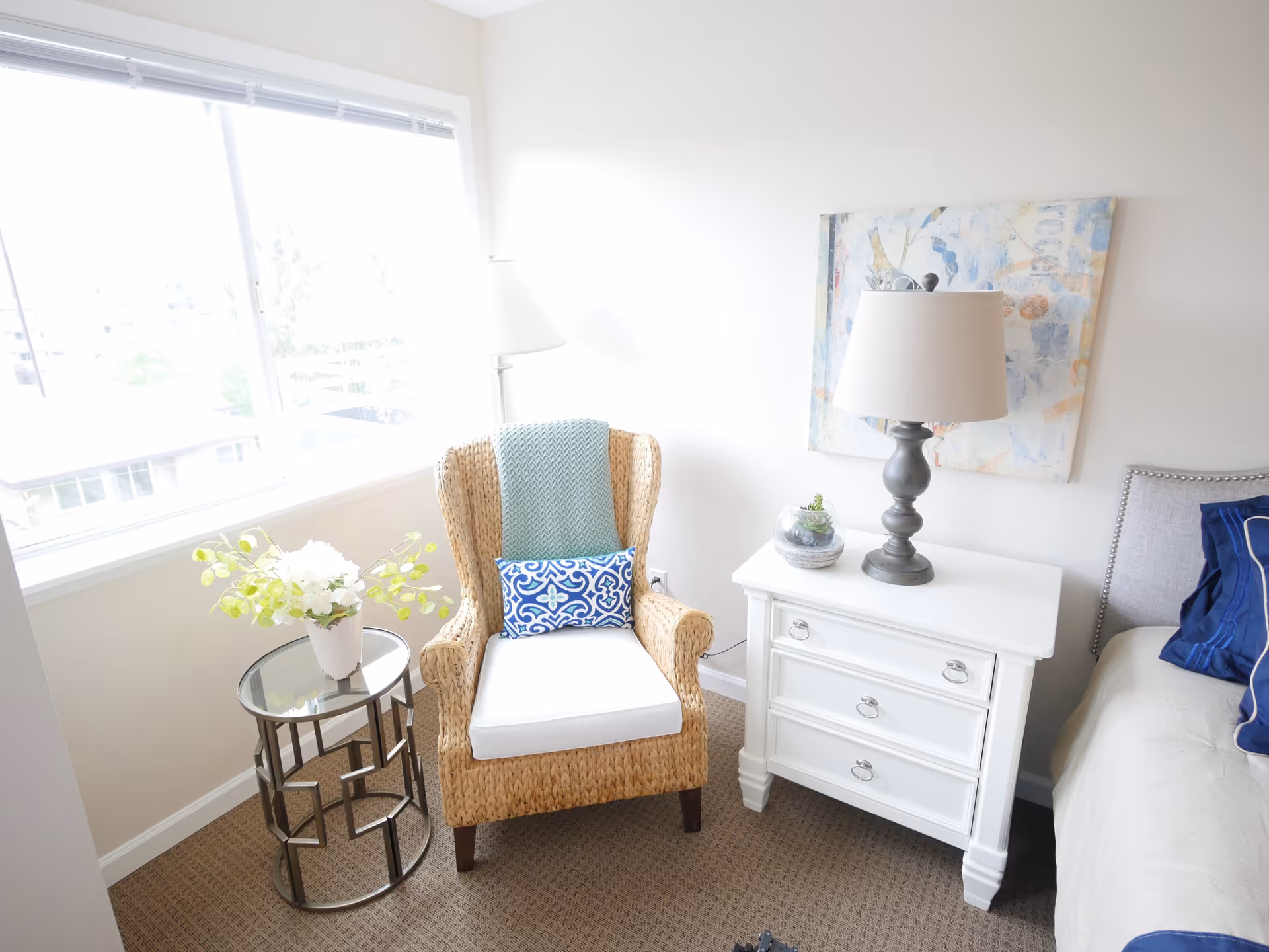 Sunlit bedroom corner with a wicker armchair, glass side table with flowers, a white nightstand topped by a lamp, and part of a bed.