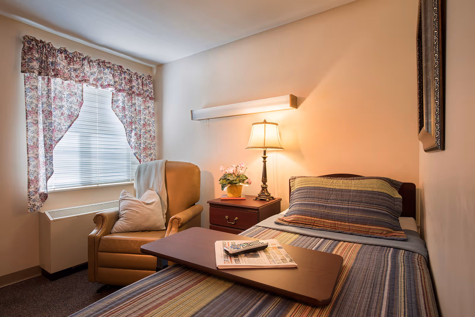 A cozy single-occupancy bedroom with a bed, bedside table and lamp, an armchair by a curtained window, and a tray table holding a newspaper and remote.