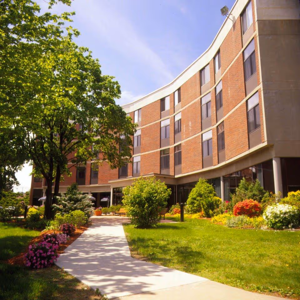 A curved multi-story brick building with large windows, surrounded by a well-maintained garden with green grass, colorful flowers, and trees under a clear blue sky.