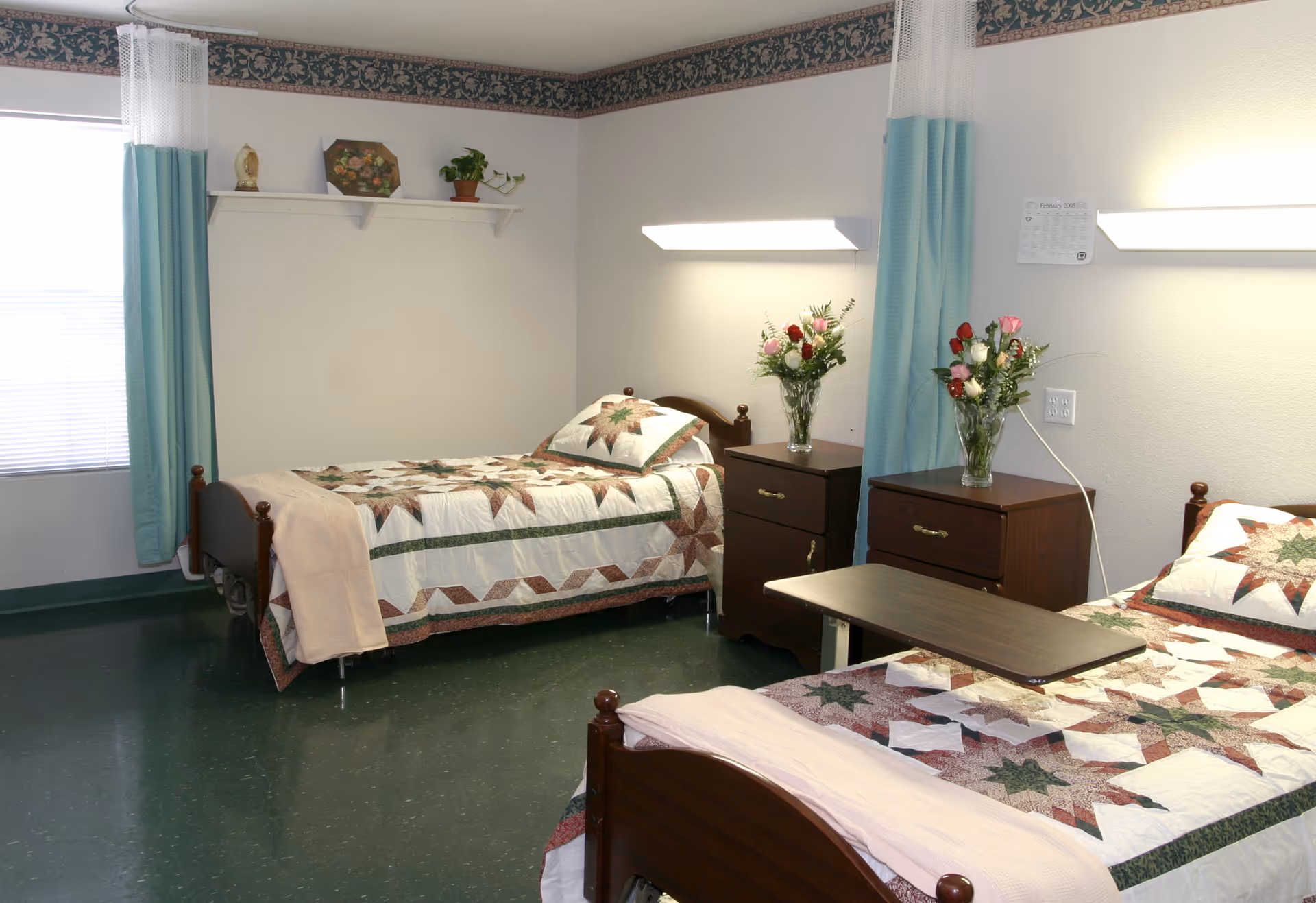 Shared nursing facility bedroom with two single beds, matching quilts, wooden nightstands with vases of flowers, curtains and overhead lights.